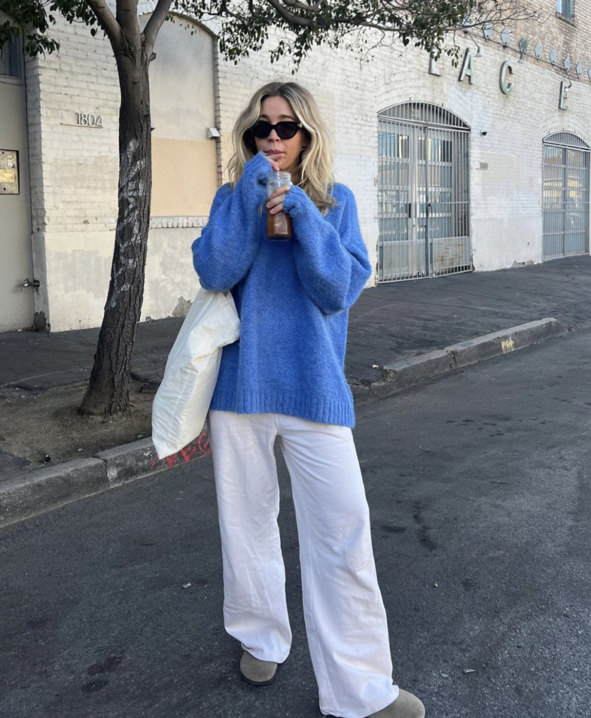 A woman enjoys a beverage on a sunny street, wearing an oversized, vibrant blue knitted sweater paired with white wide-leg pants. She accessorizes with dark sunglasses and carries a lightweight white jacket. Her blonde hair is styled in loose waves around her face, and she looks relaxed and content, with the urban environment featuring a white brick building and gated windows in the background.